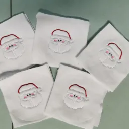 Pięć białych, złożonych kwadratów materiału z wyhaftowanym motywem Świętego Mikołaja z czerwoną czapką i różowymi policzkami, ułożonych na zielonkawym tle.