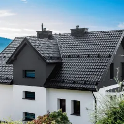 Dwukondygnacyjny dom z ciemną dachówką karpiówką, czarnymi obróbkami blacharskimi i kominami, na tle górskiego krajobrazu pod błękitnym niebem.