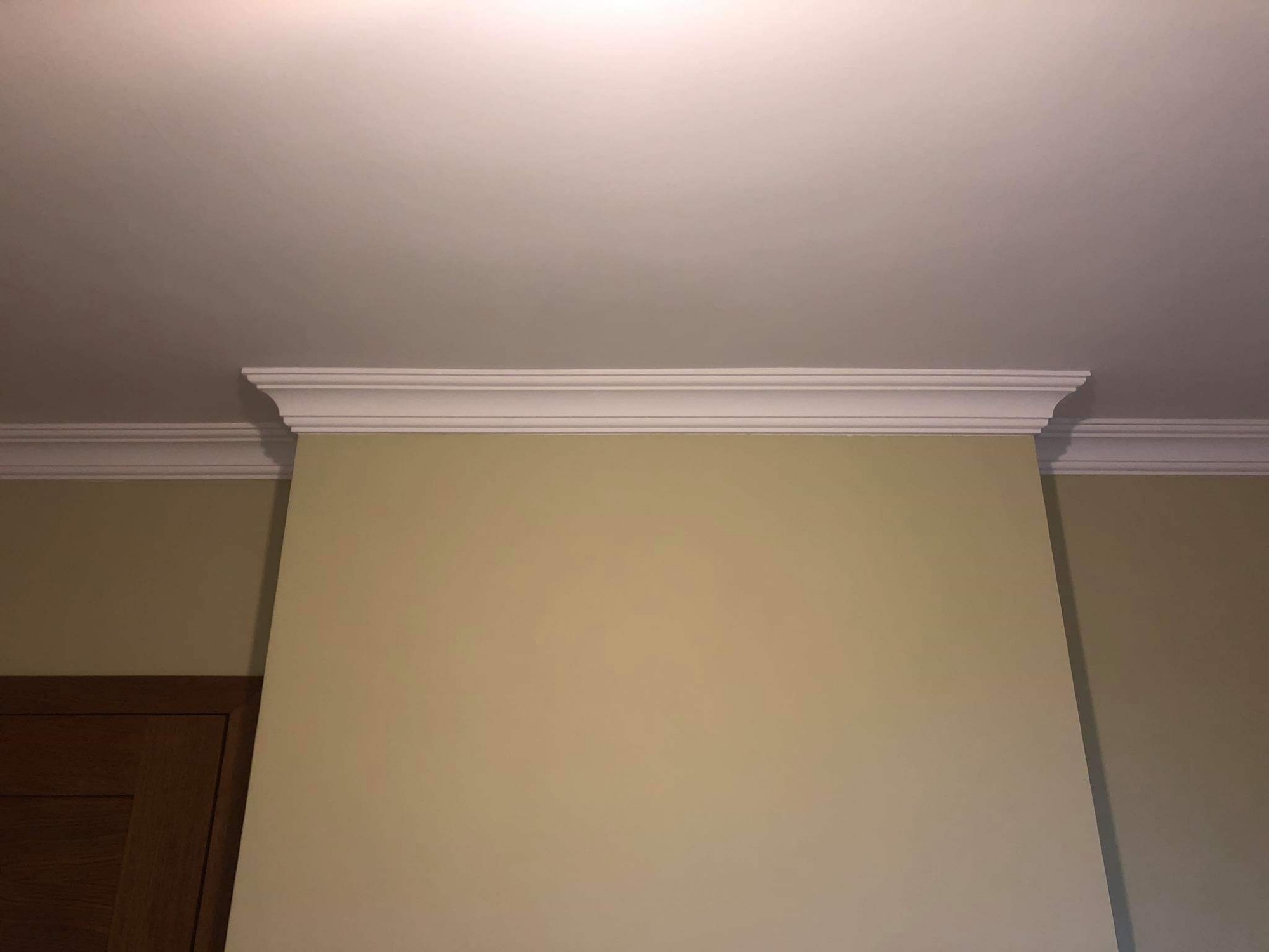 Wykończona ściana z kremowym malowaniem i dekoracyjnymi białymi listwami przysufitowymi, widoczny fragment drewnianych drzwi po lewej stronie.