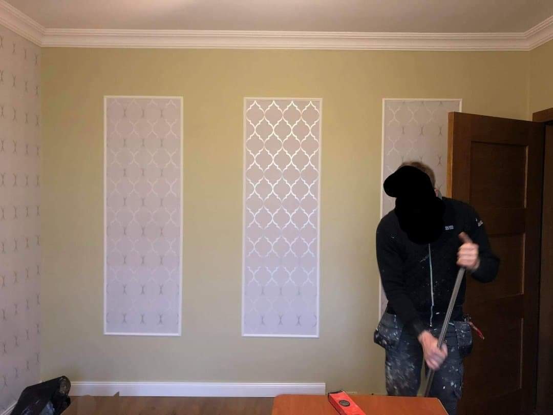 Pracownik w trakcie przygotowywania ściany do malowania dekoracyjnego z użyciem szablonów, widoczne poziome wstawki z wzorem na żółtej ścianie, listwa przypodłogowa i sufitowa, narzędzia pracy...