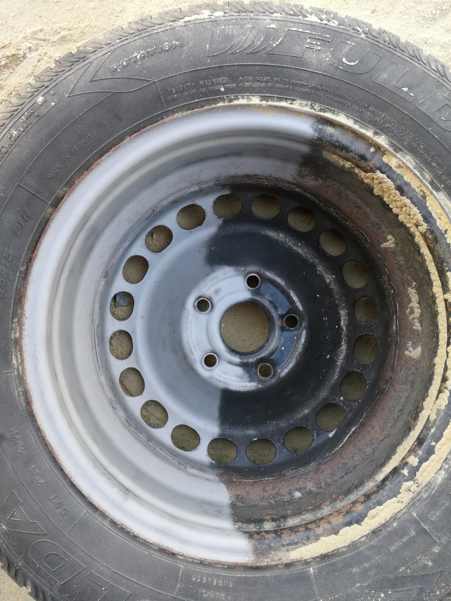 Opona samochodowa z felgą, częściowo oczyszczona z rdzy i brudu, ukazująca efekt przed i po czyszczeniu, leżąca na piasku.