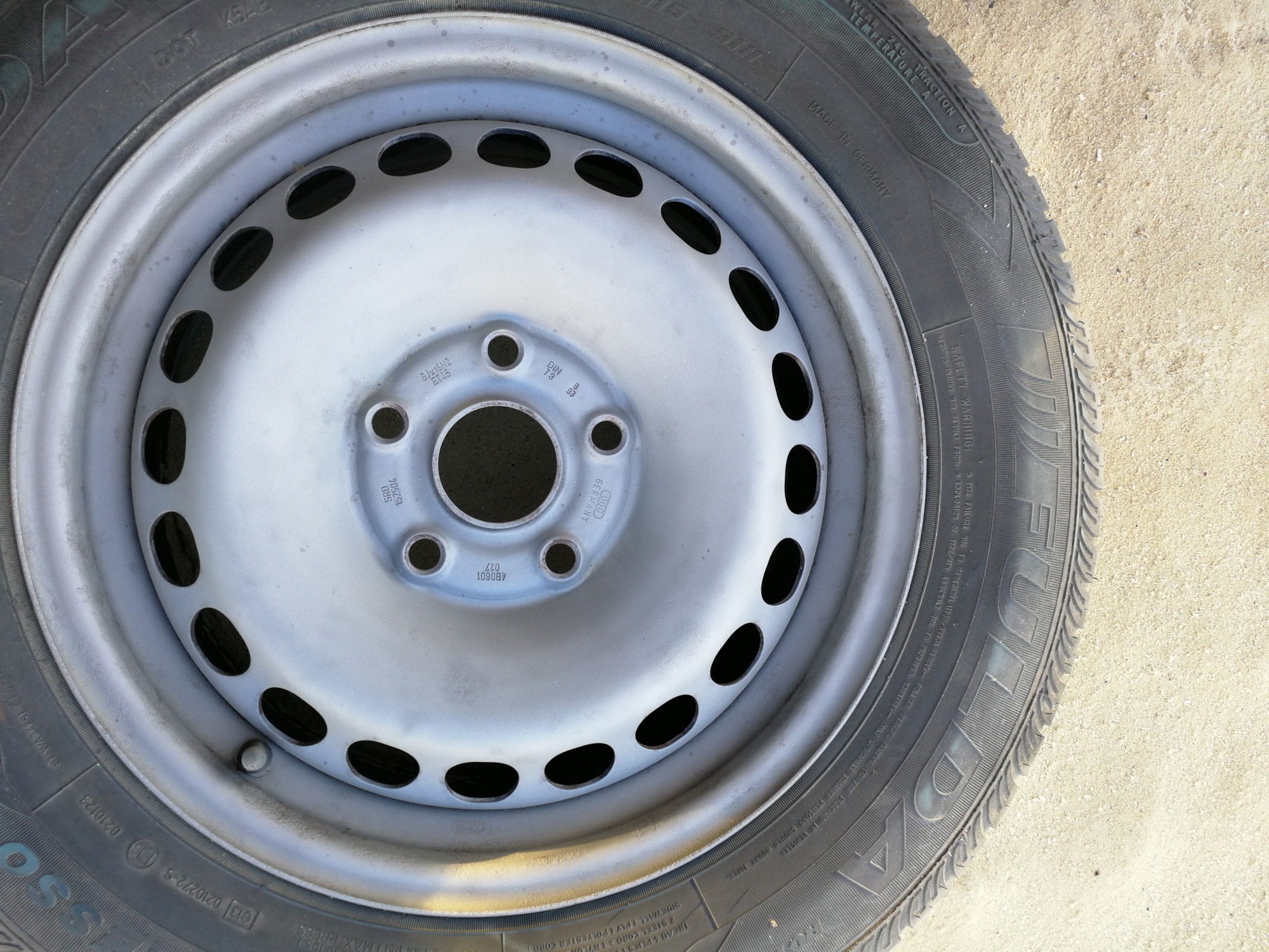 Szara stalowa felga z oponą na tle jasnego piasku, widoczne oznaczenia na oponie i feldze.