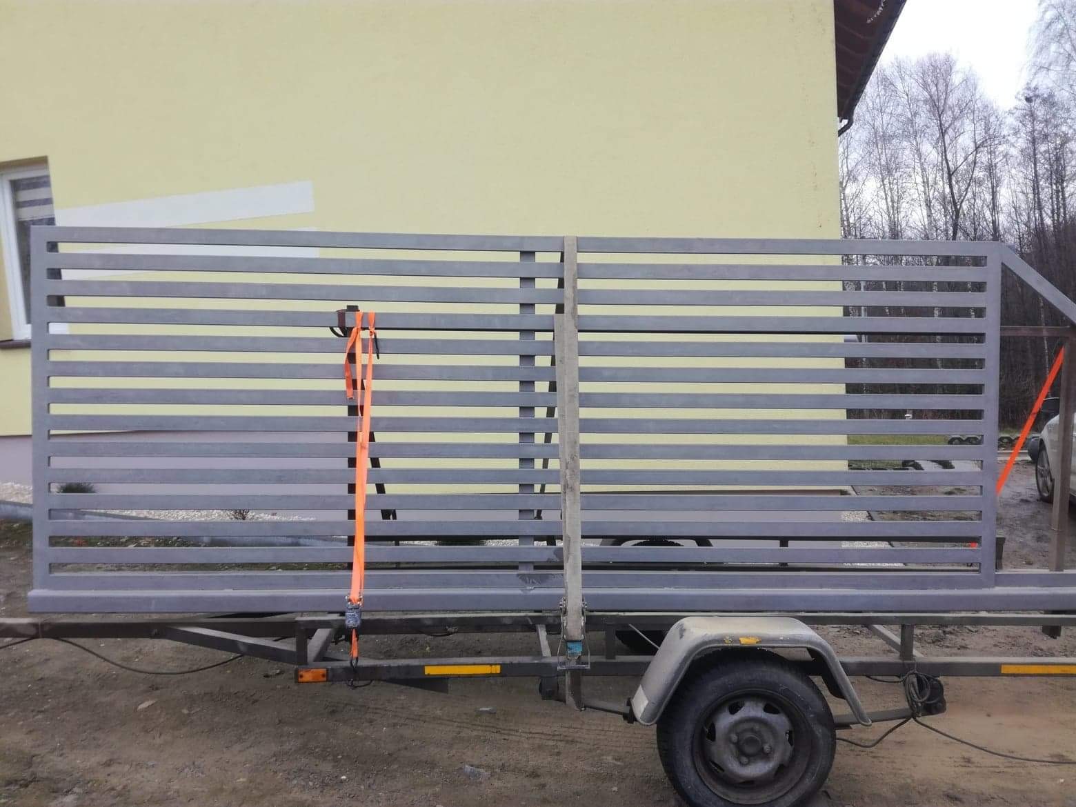 Szara, pozioma brama lamelowa, zabezpieczona pomarańczowym pasem transportowym, przewożona na przyczepce samochodowej na tle żółtej ściany budynku.