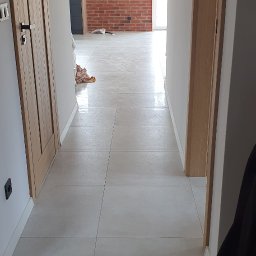 CARLOS Profesjonalne wykończenia wnętrz - Jasne, kwadratowe płytki podłogowe w korytarzu z widokiem na ścianę z czerwonej cegły w oddali, drewniane drzwi po lewej stronie i częściowo widoczny element garderoby po prawej.