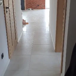 Jasne, kwadratowe płytki podłogowe w korytarzu z widokiem na ścianę z czerwonej cegły w oddali, drewniane drzwi po lewej stronie i częściowo widoczny element garderoby po prawej.