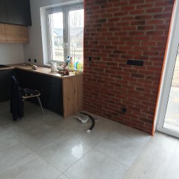 CARLOS Profesjonalne wykończenia wnętrz - Wnętrze w Słupsku z wykończoną ścianą z czerwonej cegły, zabezpieczoną pomarańczową taśmą, oraz z położonymi jasnymi płytkami podłogowymi i panelami. Widoczna część zabudowy kuchennej w kolorach...