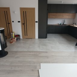 CARLOS Profesjonalne wykończenia wnętrz - Wnętrze w trakcie wykańczania: Jasnoszare płytki podłogowe przechodzą w panele podłogowe, w tle widoczna nowoczesna kuchnia z czarnymi szafkami i drewnianym blatem oraz dwoje drewnianych drzwi...