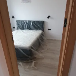 Nowe, szare łóżko z materacem w folii ochronnej, widoczne przez otwarte, drewniane drzwi w białym pokoju z jasną podłogą i nowoczesnym oświetleniem.