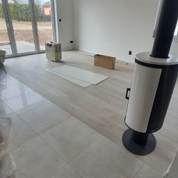 CARLOS Profesjonalne wykończenia wnętrz - Wnętrze w trakcie prac wykończeniowych: połączenie płytek podłogowych i paneli, widoczny piec wolnostojący, karton, grzejnik przenośny i płyty gipsowe