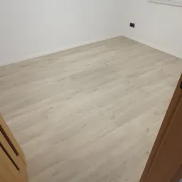 Jasne panele podłogowe ułożone w pokoju z białymi ścianami, widoczny fragment ościeżnicy drewnianej i szara płytka przy progu.