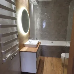 Nowoczesna łazienka z drewnopodobną podłogą, szarą glazurą, podświetlanym lustrem owalnym nad białą szafką z drewnianym blatem i grzejnikiem drabinkowym po lewej stronie.