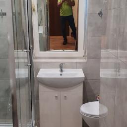 CARLOS Profesjonalne wykończenia wnętrz - Ukończona łazienka z kabiną prysznicową, umywalką z szafką i toaletą, widoczne szare płytki na ścianach i drewnopodobne panele na podłodze, w lustrze odbija się osoba robiąca zdjęcie.