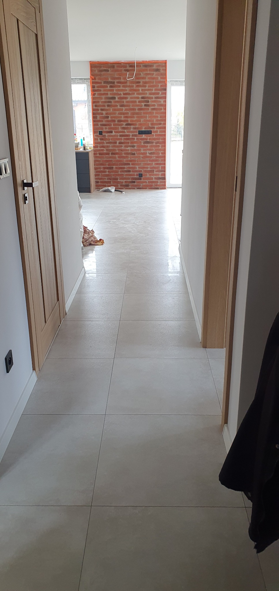 Jasne, kwadratowe płytki podłogowe w korytarzu z widokiem na ścianę z czerwonej cegły w oddali, drewniane drzwi po lewej stronie i częściowo widoczny element garderoby po prawej.