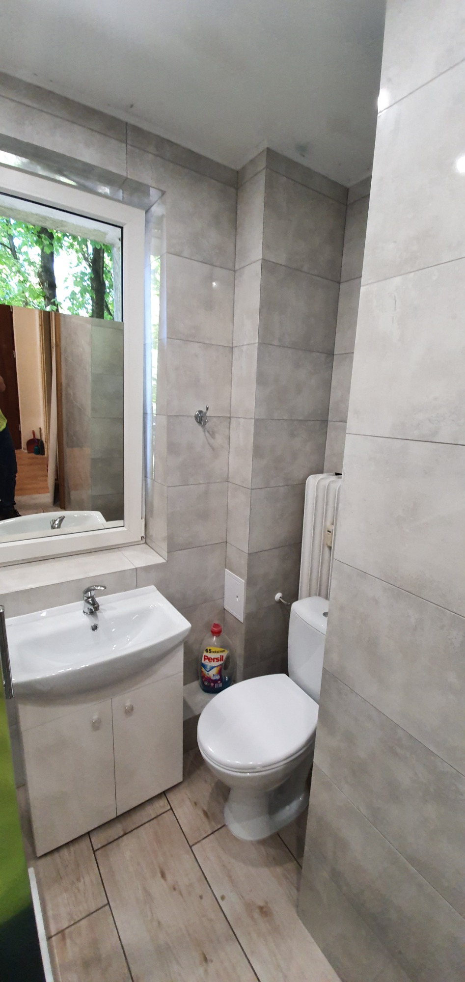 Wykończona łazienka z szarymi płytkami, białą umywalką z szafką, toaletą i grzejnikiem, widok z perspektywy drzwi, z butelką płynu do prania na podłodze.