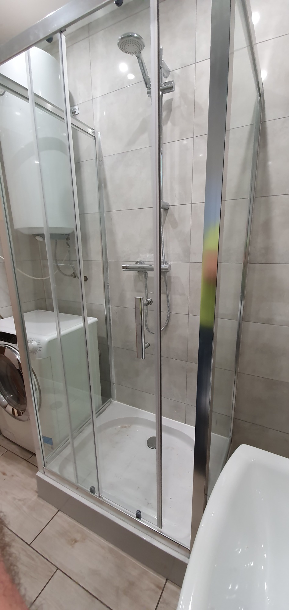 Szklana kabina prysznicowa z brodzikiem, widoczna bateria, deszczownica i pralka w tle, szare płytki na ścianie.