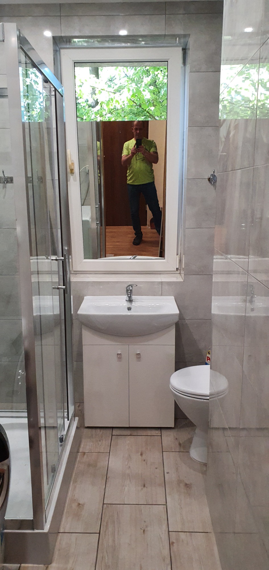 Ukończona łazienka z kabiną prysznicową, umywalką z szafką i toaletą, widoczne szare płytki na ścianach i drewnopodobne panele na podłodze, w lustrze odbija się osoba robiąca zdjęcie.