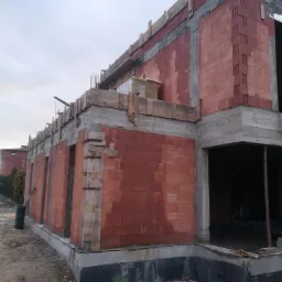 Surowy stan budynku w trakcie budowy, widoczne czerwone bloczki ceramiczne, szalunki i zbrojenia, zarys przyszłych okien i drzwi, teren budowy z ziemią i fragmentem rury kanalizacyjnej.