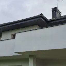 Fragment domu z ciemnym dachem, kominem z anteną i jasną elewacją w pochmurny dzień.