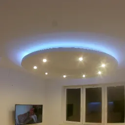 Okrągły podwieszany sufit z oświetleniem LED i punktowymi lampami w nowoczesnym salonie z białymi ścianami i oknem.