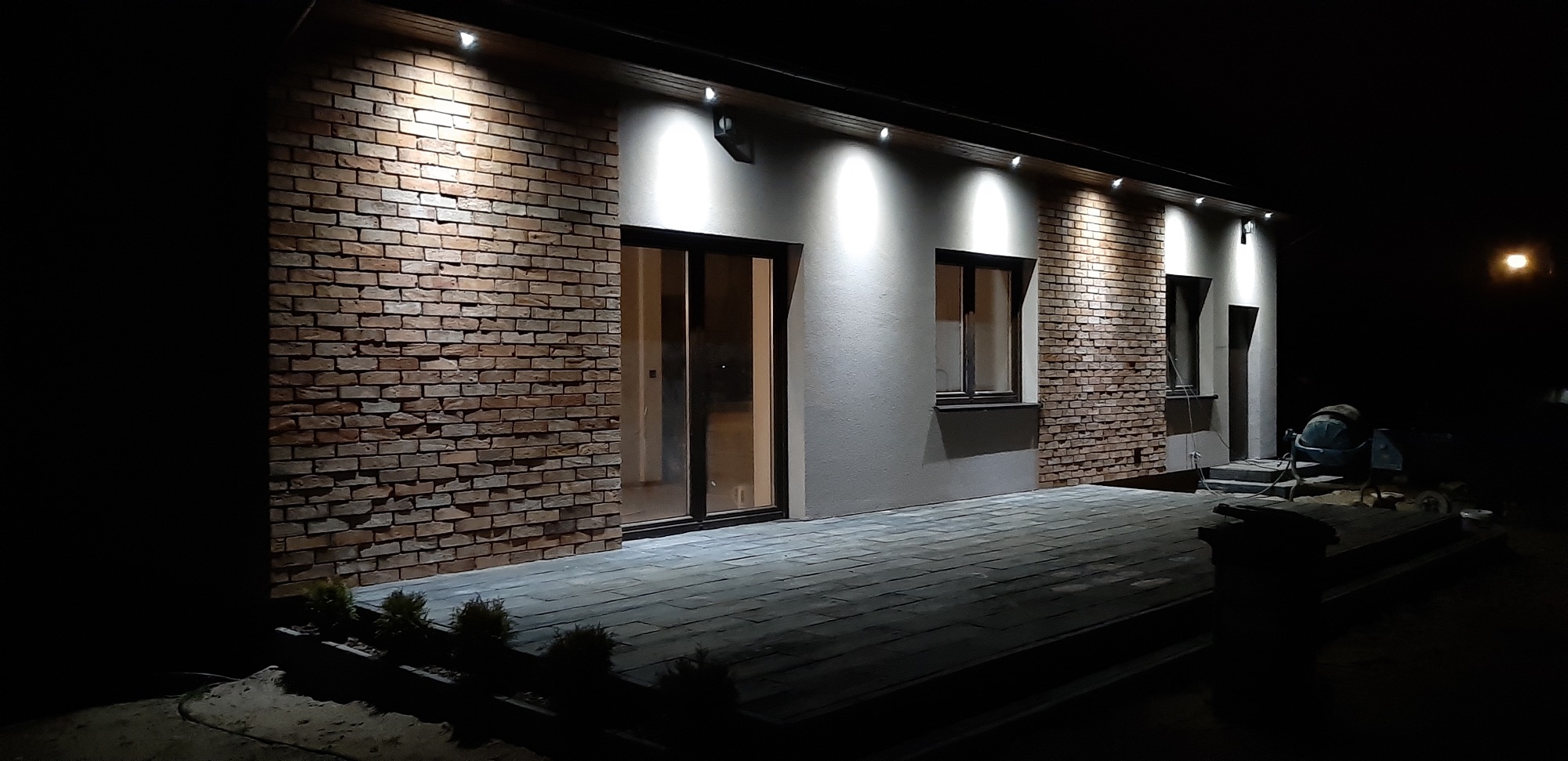 Nocne ujęcie wykończonej elewacji domu z cegły klinkierowej i jasnego tynku, oświetlonej punktowymi lampami elewacyjnymi; widoczny taras z kostki brukowej i betoniarka w tle.