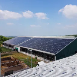 Instalacja paneli słonecznych na dachu budynku gospodarczego, widok z góry na fragment dachu z konstrukcją montażową w tle budynek z panelami i błękitne niebo z chmurami.