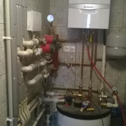 Gdak Grażyna Eko Instal - Kotłownia z białym kotłem gazowym Vaillant, zbiornikiem, czerwonymi pompami obiegowymi i licznymi rurami w pomieszczeniu wyłożonym szarą, wzorzystą płytką.