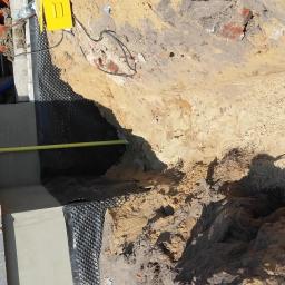 Gdak Grażyna Eko Instal - Fundament budynku w trakcie izolacji: ściana z bloczków betonowych, izolacja przeciwwilgociowa z folii kubełkowej, wystająca rura gazowa, wykop z ziemią.