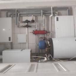 Gdak Grażyna Eko Instal - Wnętrze kotłowni z zainstalowanym bojlerem, zbiornikiem wyrównawczym, pompami obiegowymi i orurowaniem na tle płytek ceramicznych.