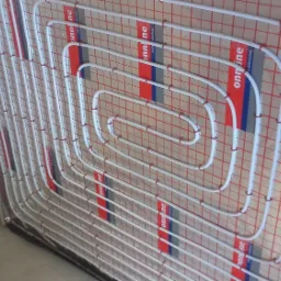 Białe rury grzewcze ułożone spiralnie na czerwonej siatce mocującej, widok z boku na podłogę w trakcie instalacji.