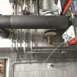 Skomplikowany system rur i zaworów w kotłowni, widoczny czarny przewód kominowy, automatyka kotła oraz rozdzielacz ogrzewania podłogowego z czerwonymi pętlami.