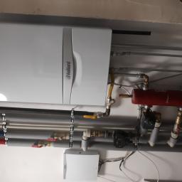 Gdak Grażyna Eko Instal - Montaż kotła grzewczego marki Vaillant wraz z osprzętem i orurowaniem w Grodzisku Mazowieckim. Widoczne liczne rury, zawory, zbiornik wyrównawczy oraz regulator temperatury.