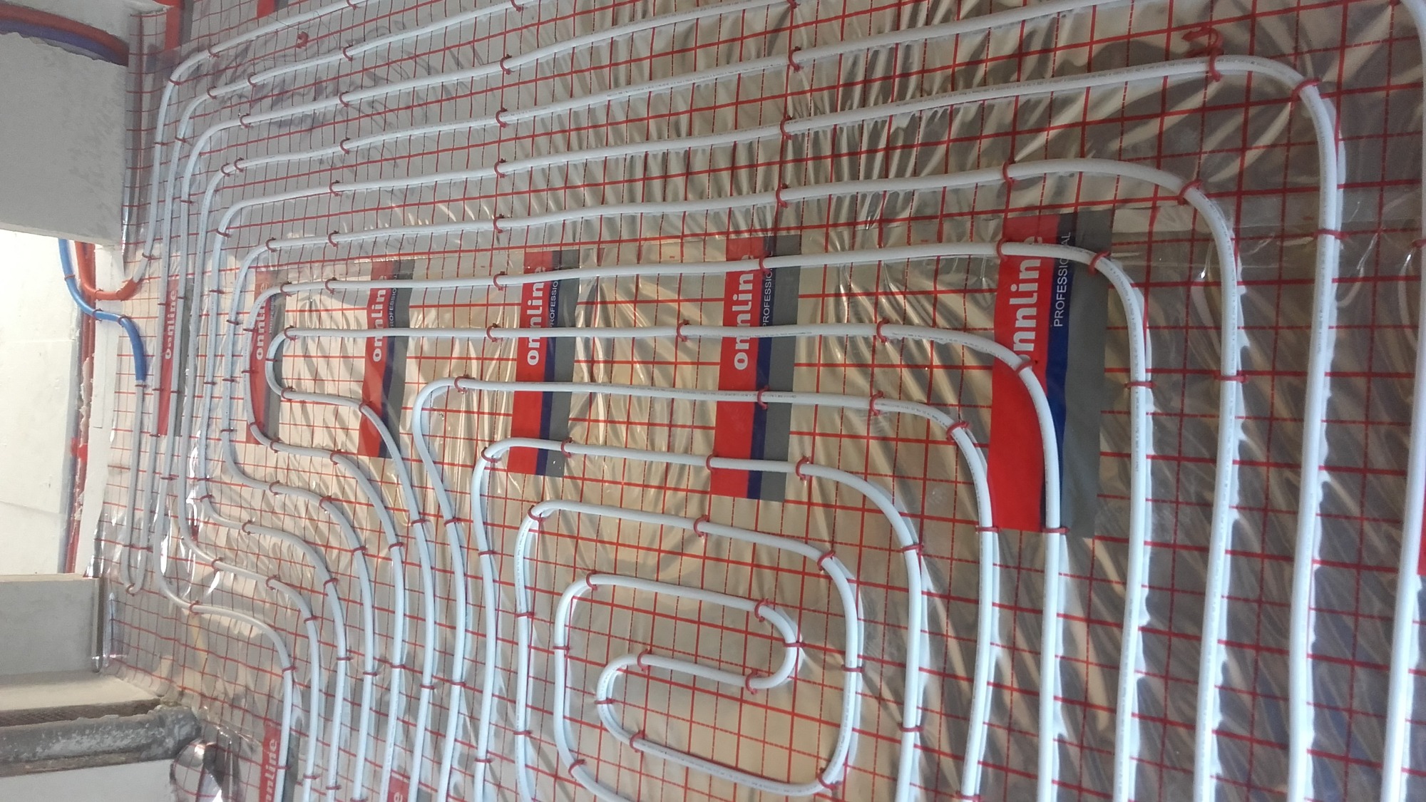 Układ białych rur ogrzewania podłogowego zamocowanych czerwonymi klipsami na folii z nadrukowaną czerwoną siatką, widoczne fragmenty ściany i podłogi.