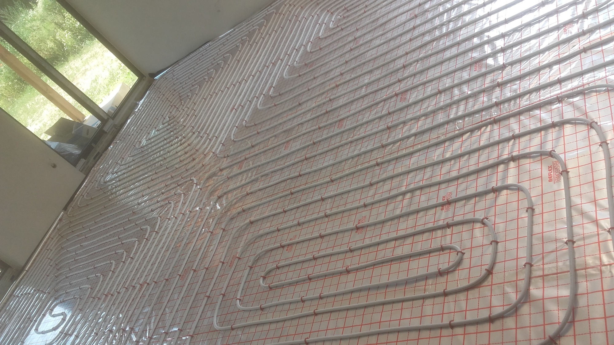 Białe rurki ogrzewania podłogowego ułożone gęsto na izolacji z czerwoną siatką, widok z boku, w tle okno.