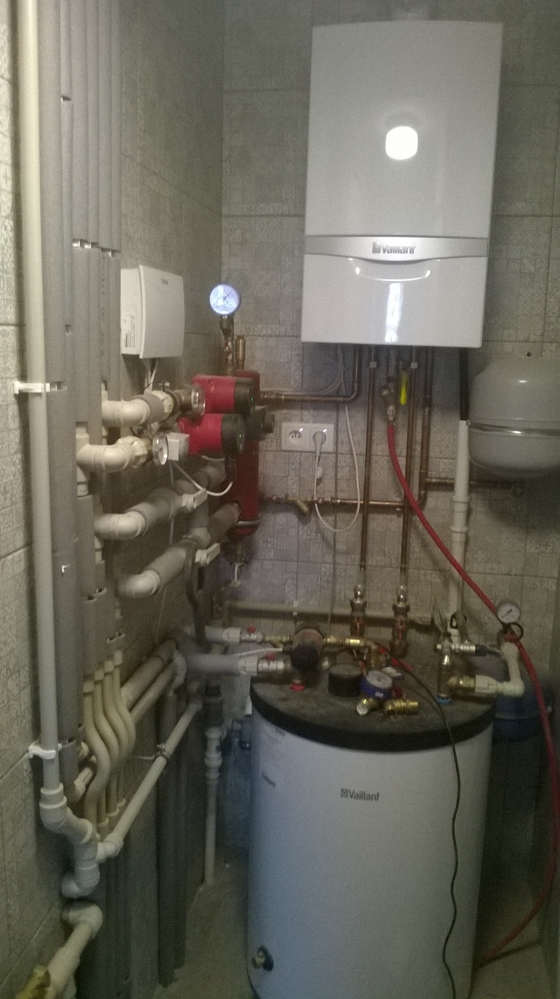 Kotłownia z białym kotłem gazowym Vaillant, zbiornikiem, czerwonymi pompami obiegowymi i licznymi rurami w pomieszczeniu wyłożonym szarą, wzorzystą płytką.
