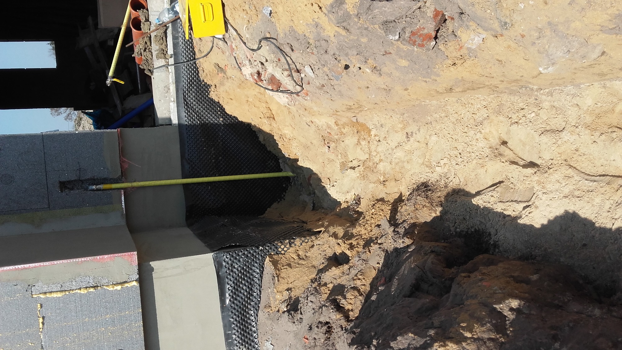 Fundament budynku w trakcie izolacji: ściana z bloczków betonowych, izolacja przeciwwilgociowa z folii kubełkowej, wystająca rura gazowa, wykop z ziemią.