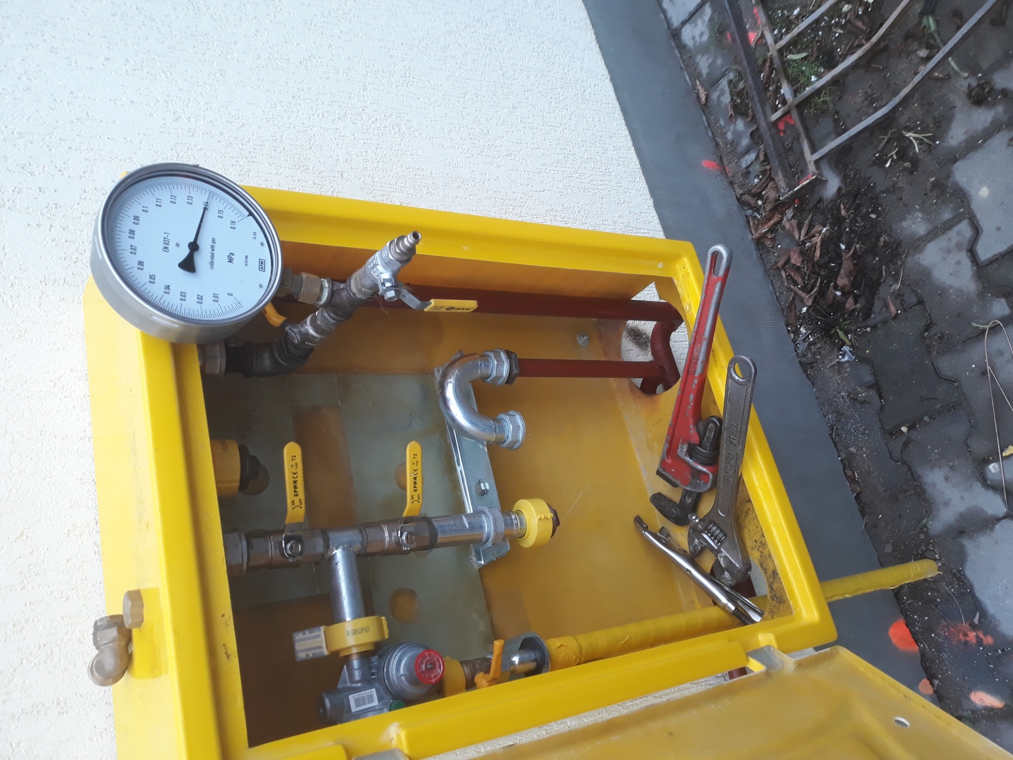 Żółta skrzynka gazowa z manometrem, zaworami i rurami, widoczne narzędzia: klucz nastawny i żabka