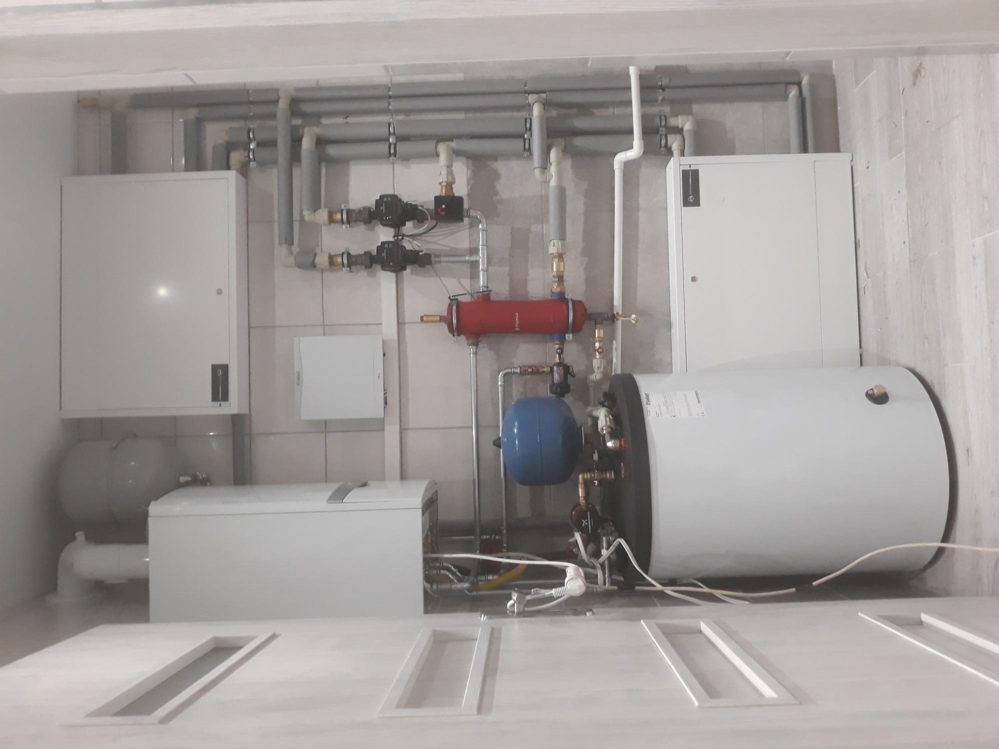 Wnętrze kotłowni z zainstalowanym bojlerem, zbiornikiem wyrównawczym, pompami obiegowymi i orurowaniem na tle płytek ceramicznych.