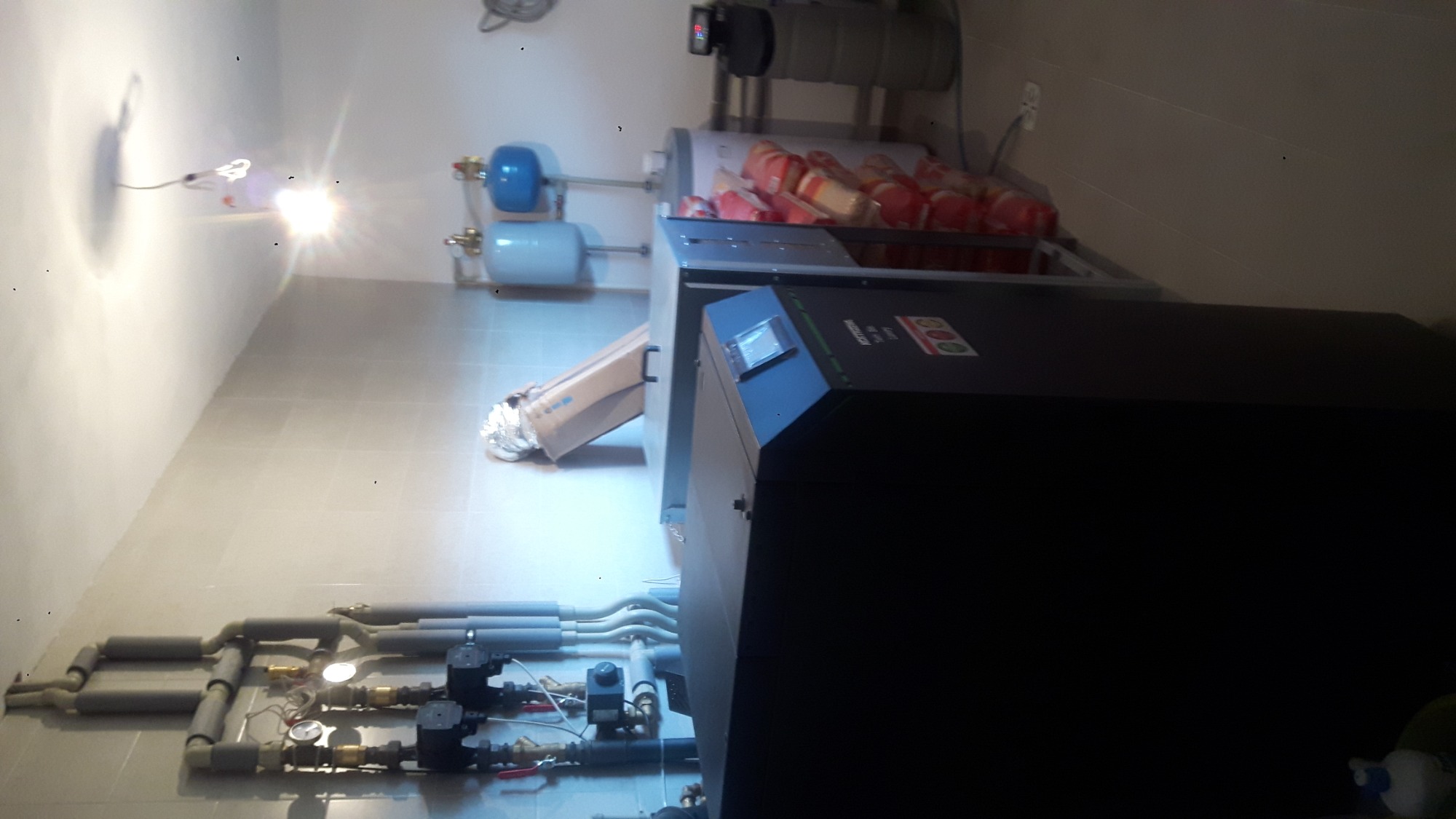 Kotłownia z nowoczesnym piecem, zbiornikami wyrównawczymi, izolowanymi rurami i elementami instalacji, widoczne z boku, w pomieszczeniu z białymi płytkami.