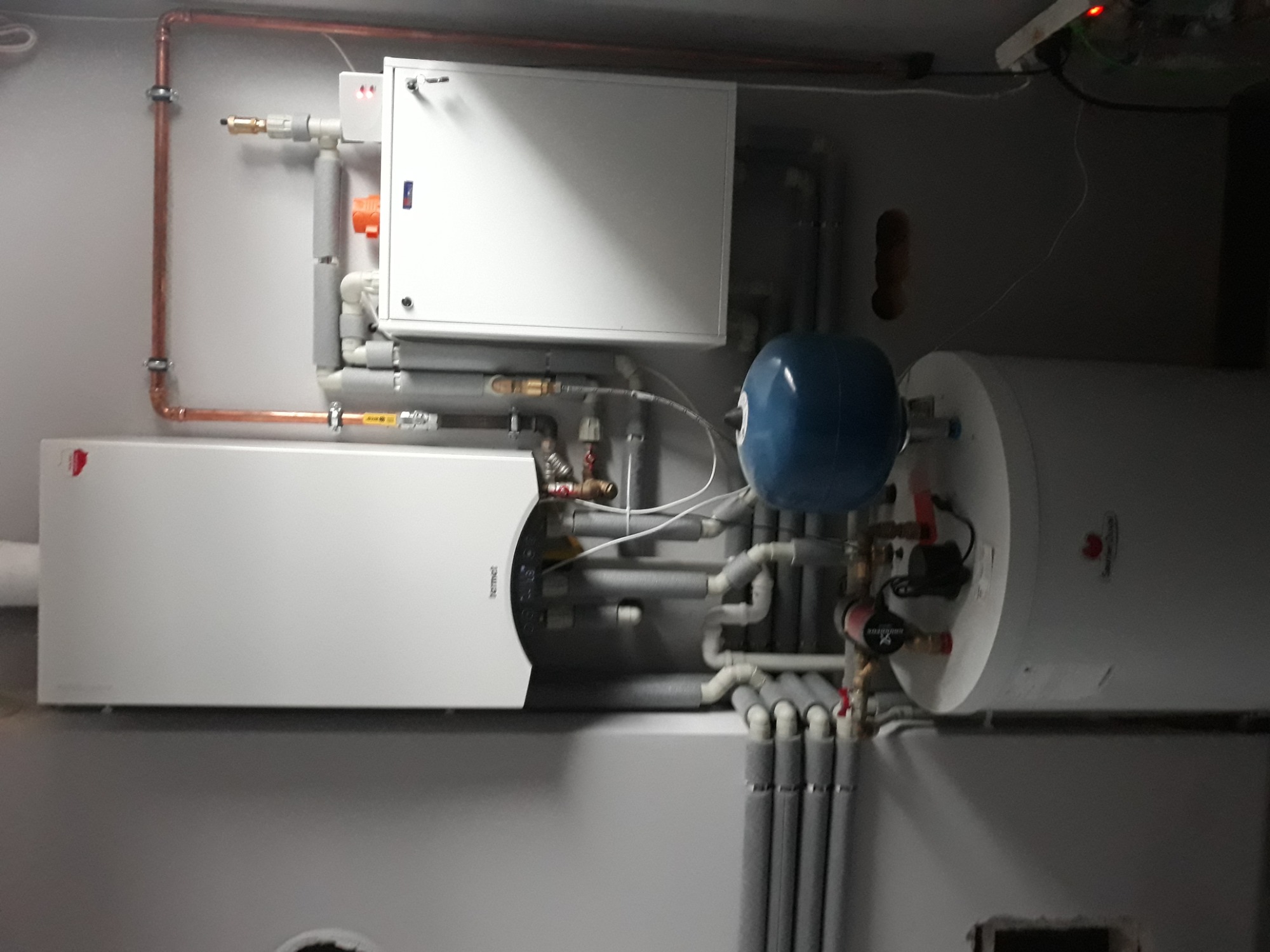 Kotłownia z białym piecem grzewczym, zasobnikiem ciepłej wody i niebieskim naczyniem przeponowym, liczne rury w izolacji termicznej, instalacja połączona miedzianymi rurami.