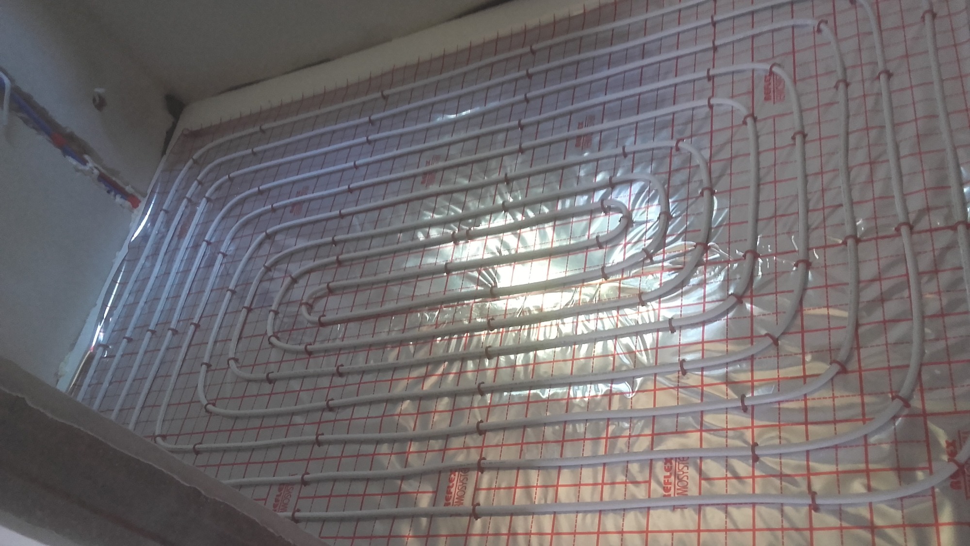 Białe rurki ogrzewania podłogowego ułożone na folii z czerwoną kratką, widoczne fragmenty instalacji hydraulicznej przy ścianie.