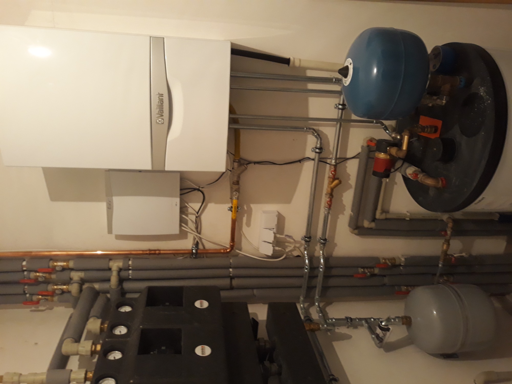 Kotłownia: biały piec gazowy Vaillant, zbiornik wyrównawczy, liczne rury miedziane i szare izolowane, manometry na czarnym urządzeniu.