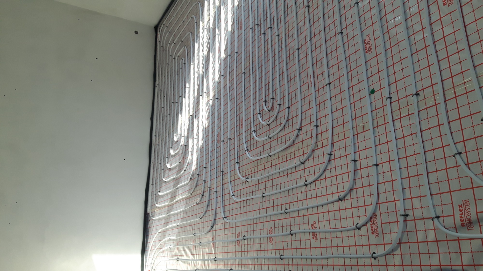 Białe rurki ogrzewania podłogowego ułożone na czerwonej kratownicy z folii izolacyjnej, mocowane czarnymi klipsami, widok z boku z perspektywą.