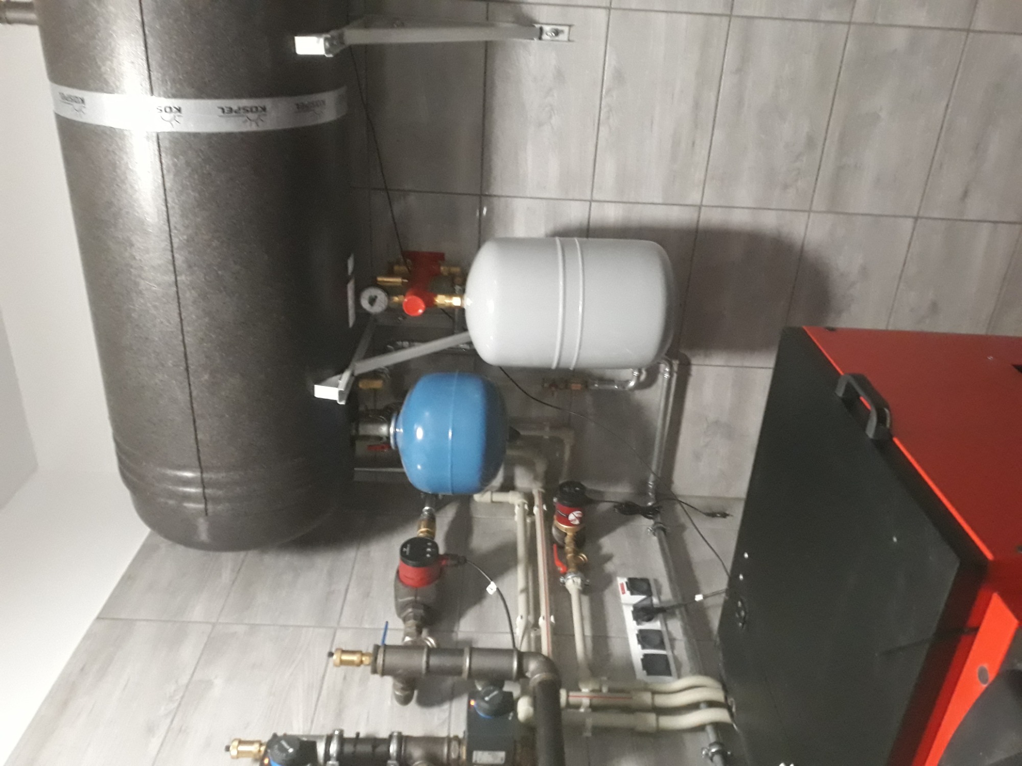 Kotłownia z grzejnikiem, zbiornikiem akumulacyjnym Kospel, naczyniami przeponowymi i orurowaniem na tle płytek ceramicznych.