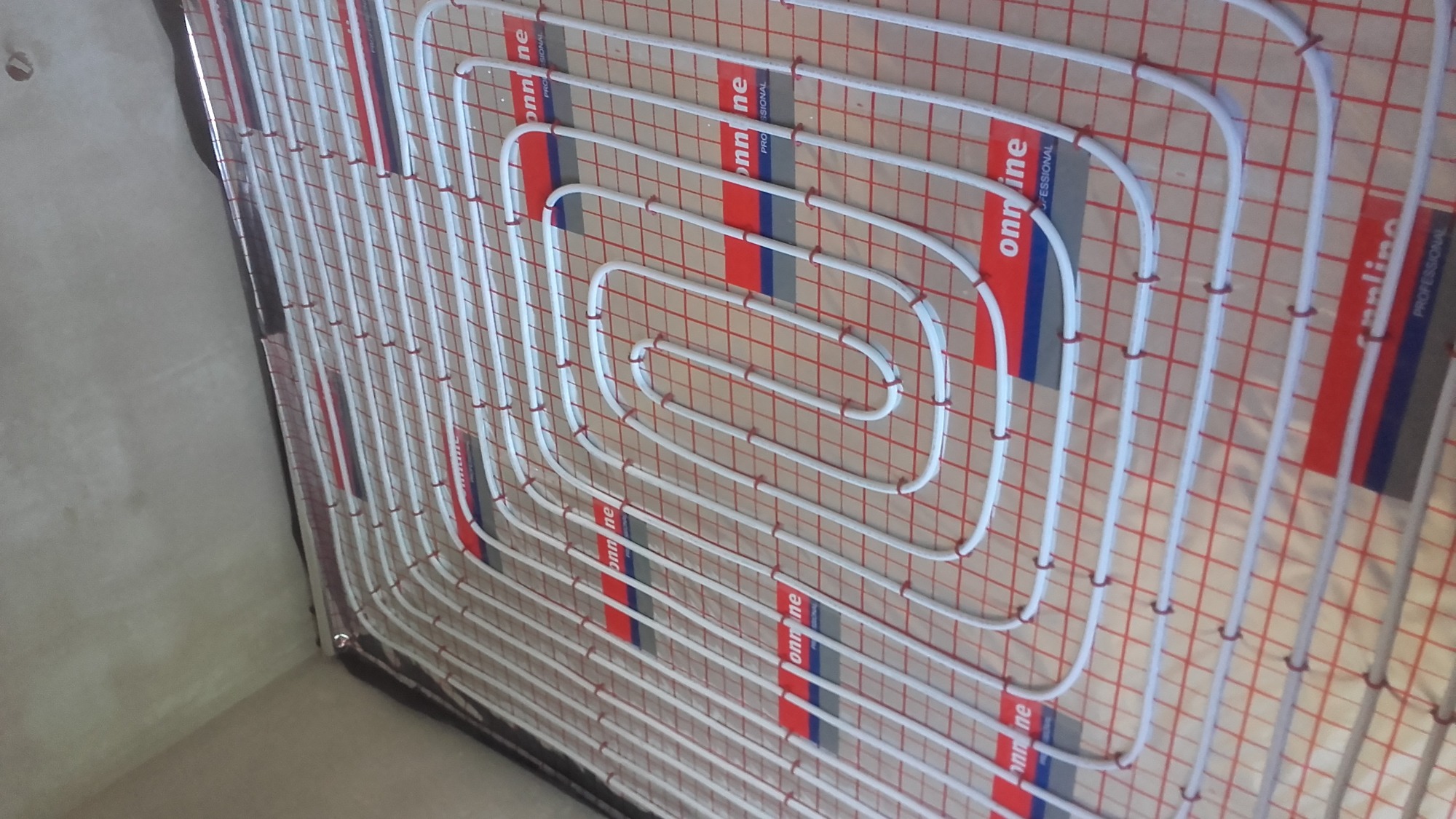 Białe rury grzewcze ułożone spiralnie na czerwonej siatce mocującej, widok z boku na podłogę w trakcie instalacji.