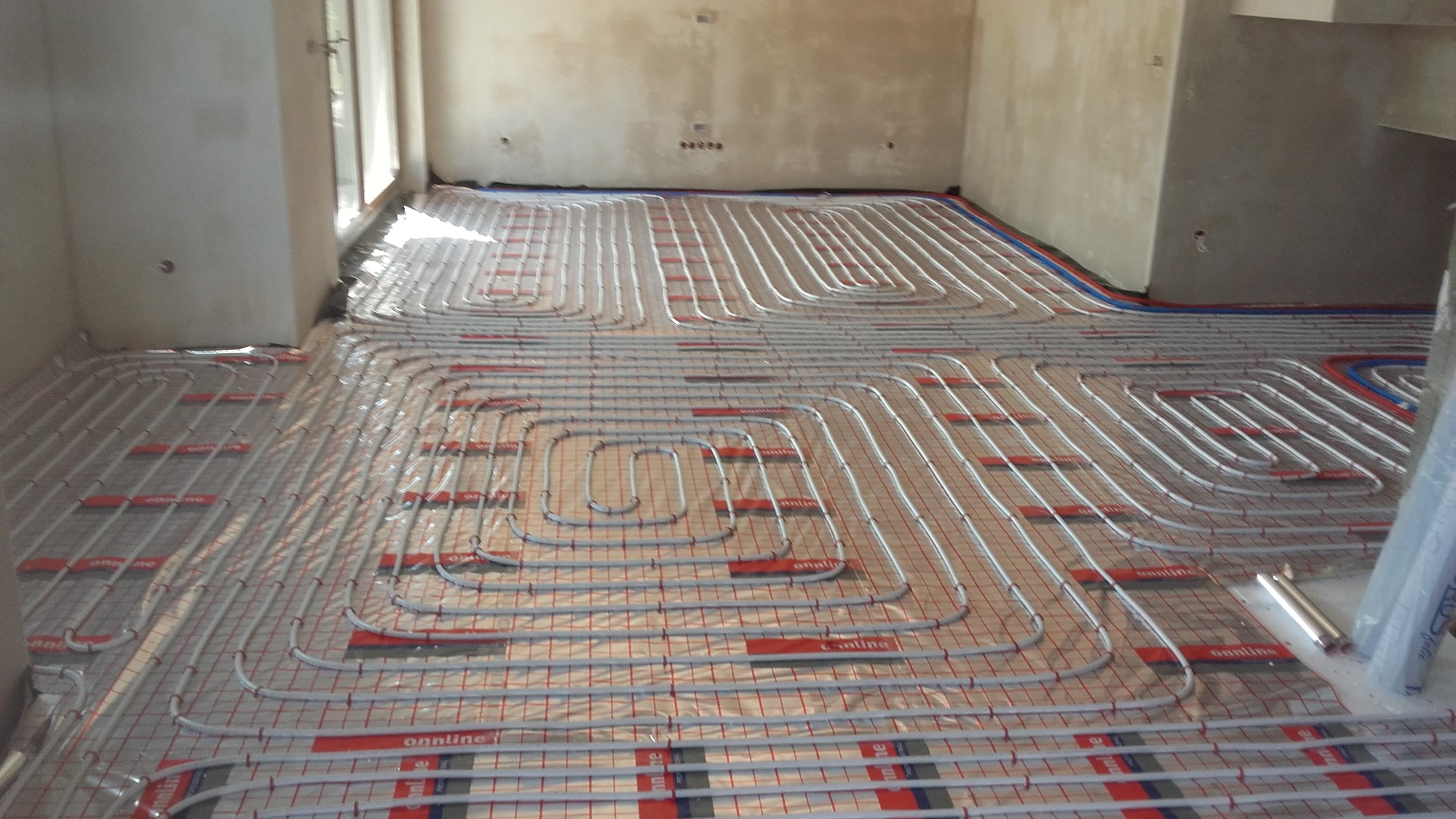 Montaż rur grzewczych podłogowych na siatce w surowym pomieszczeniu, widoczne złącza i elementy mocujące, przygotowanie do zalania wylewką.