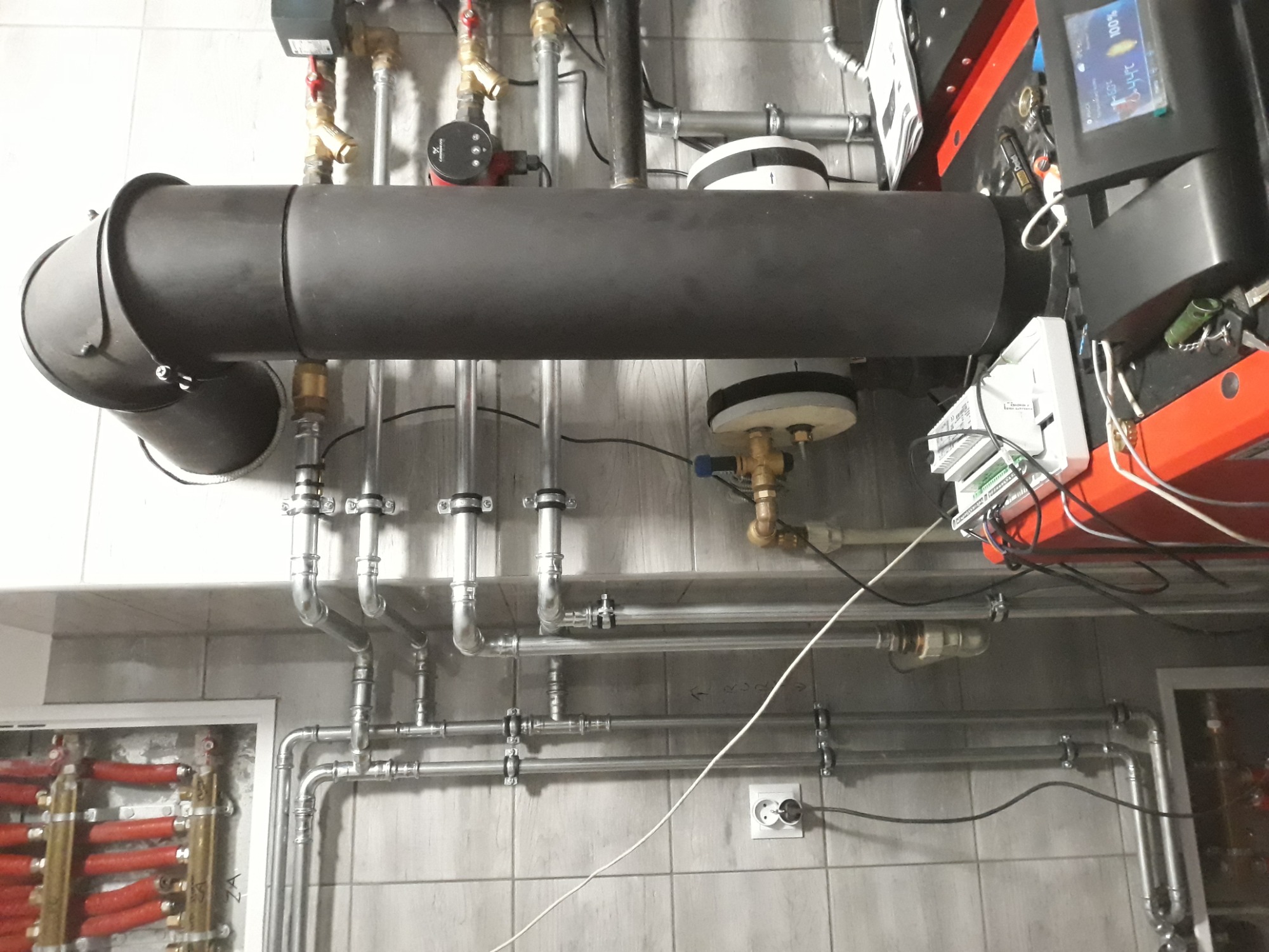 Skomplikowany system rur i zaworów w kotłowni, widoczny czarny przewód kominowy, automatyka kotła oraz rozdzielacz ogrzewania podłogowego z czerwonymi pętlami.