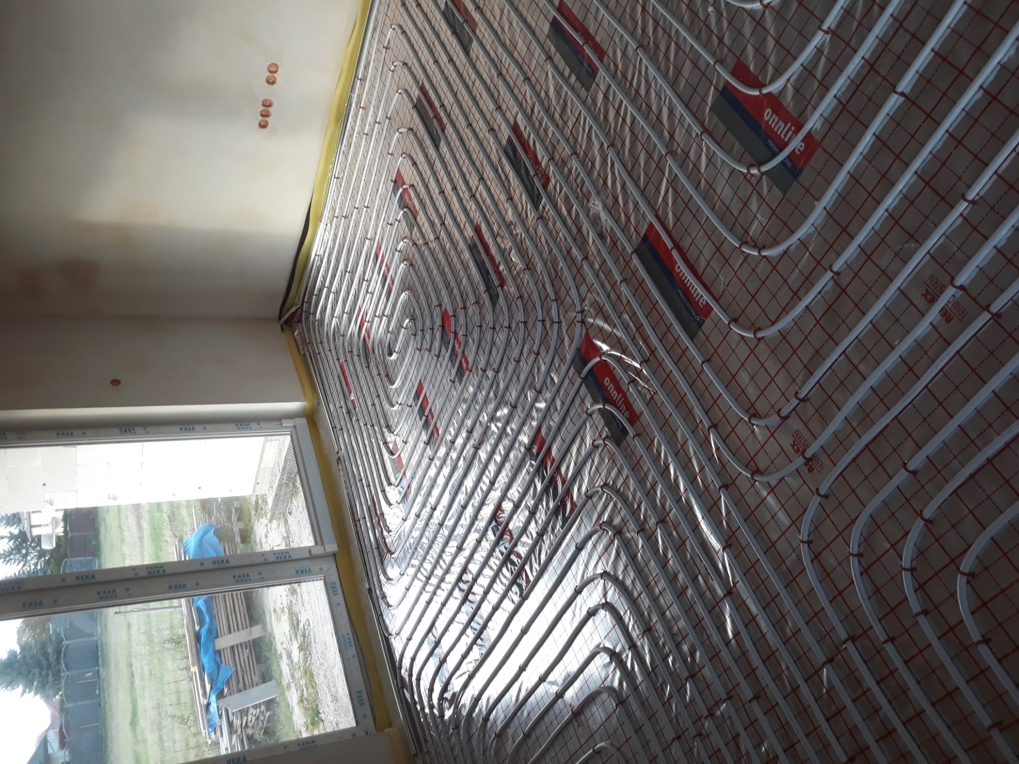 Białe rurki ogrzewania podłogowego ułożone spiralnie na folii z czerwoną kratką, widoczne okno z profilem Veka, surowy stan wykończenia wnętrza.