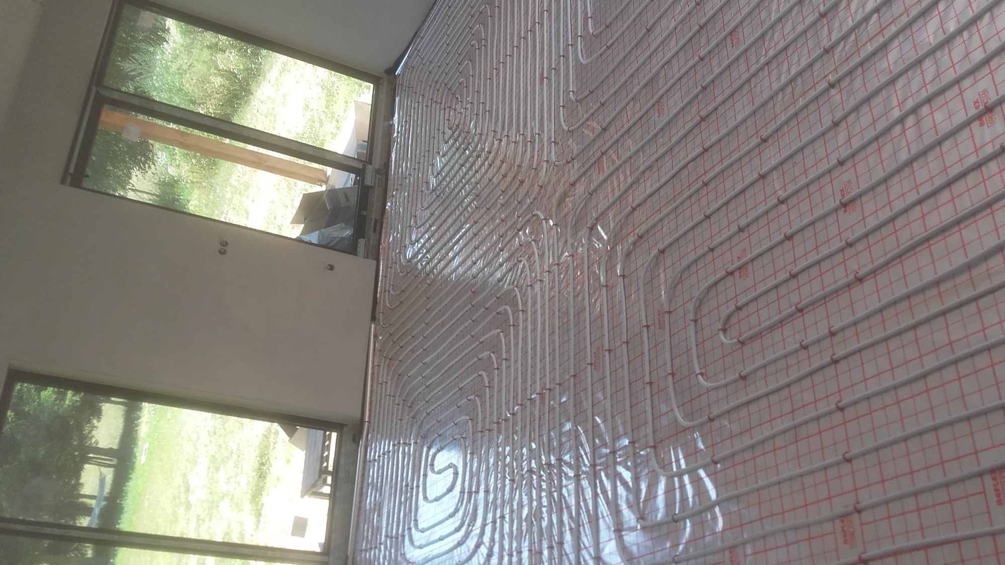 Białe rury ogrzewania podłogowego ułożone na folii z czerwoną siatką, widok z boku, okno z widokiem na zieleń w tle.