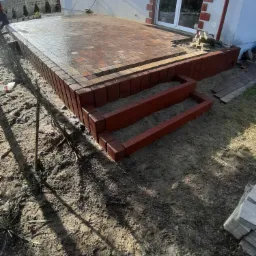 Nowo ułożony taras z kostki brukowej z dwoma stopniami wejściowymi, widoczne świeże ślady po pracy i stos kostki obok.