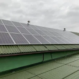 Instalacja paneli słonecznych na zielonym, falistym dachu z blachy, widoczne przewody elektryczne i wentylatory dachowe przy pochmurnym niebie.